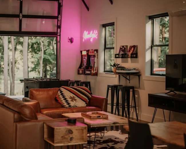 The image shows a modern living room with a leather sofa, a wooden coffee table, bar stools, and a large window revealing trees outside.