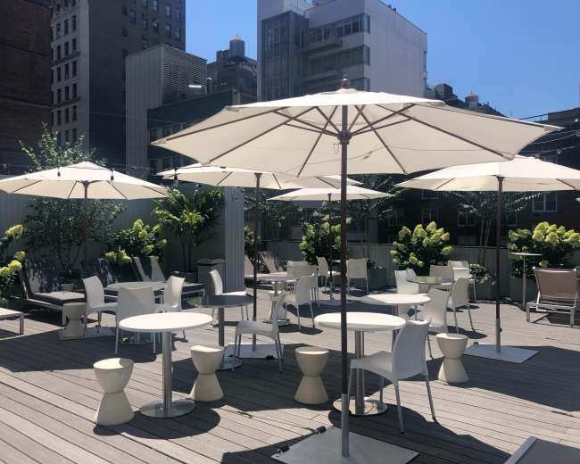 A rooftop space features white umbrellas, tables, and chairs surrounded by greenery and buildings under a clear blue sky.
