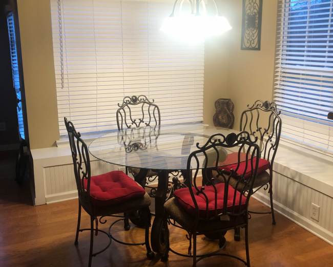 A round glass dining table with four iron chairs, each featuring red cushions, is positioned by a window in a well-lit room.