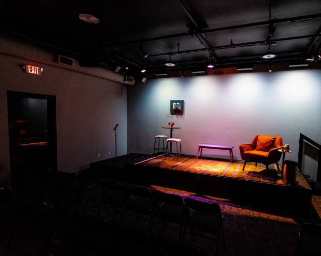 The image shows a small theater stage with a red armchair, a small table with a vase, and two stools, surrounded by empty folding chairs facing the stage.