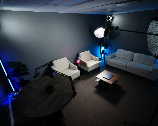 The image shows a corner of a modern, minimalistic lounge area with two white armchairs and a white sofa arranged around a coffee table, illuminated by blue and soft white lighting.