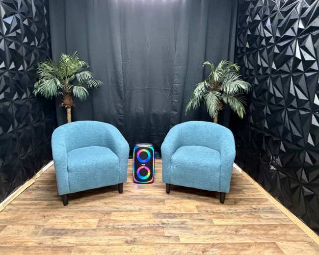 The image shows two blue chairs positioned opposite each other with two artificial palm trees and a colorful speaker in between, against a dark, textured backdrop.