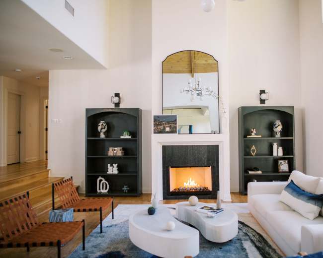 The image shows a modern living room featuring a white sofa, two brown chairs, a coffee table, and a fireplace, with large windows allowing natural light.