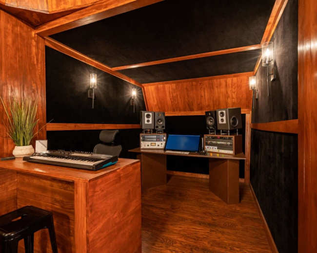 The image shows a compact music production studio with wooden walls, a central desk containing audio equipment, a keyboard, and studio monitors.
