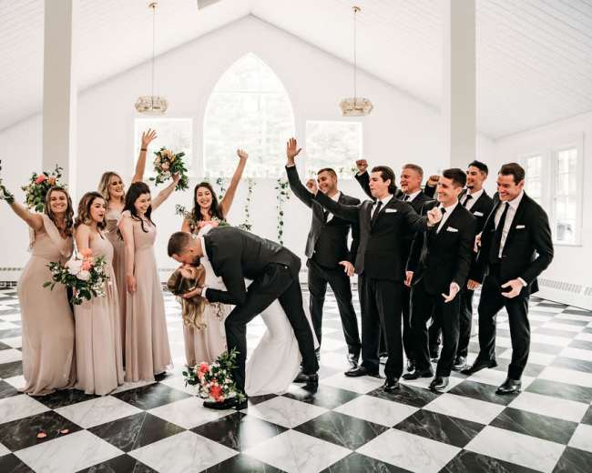 A wedding party celebrates in a spacious hall with a checkered floor and large windows, as a couple shares a kiss at the center.