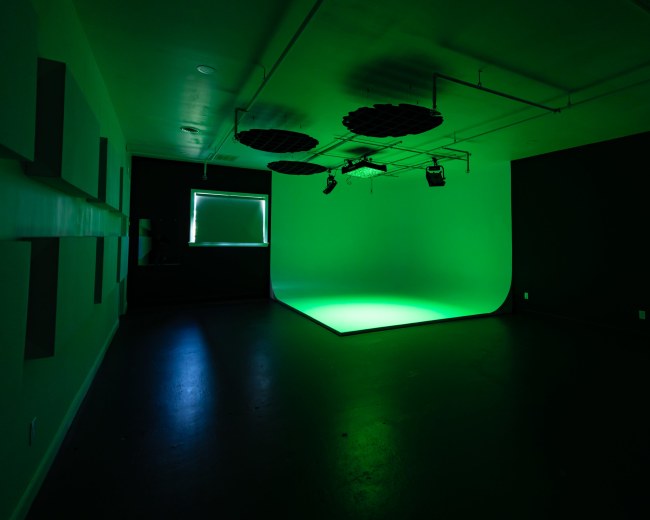 The image shows a studio space with green lighting and a curved backdrop, featuring dark walls and overhead lighting fixtures.