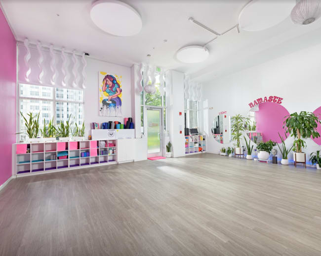 The image shows a spacious yoga studio with pink accent walls, plants, and exercise equipment neatly organized on shelves.