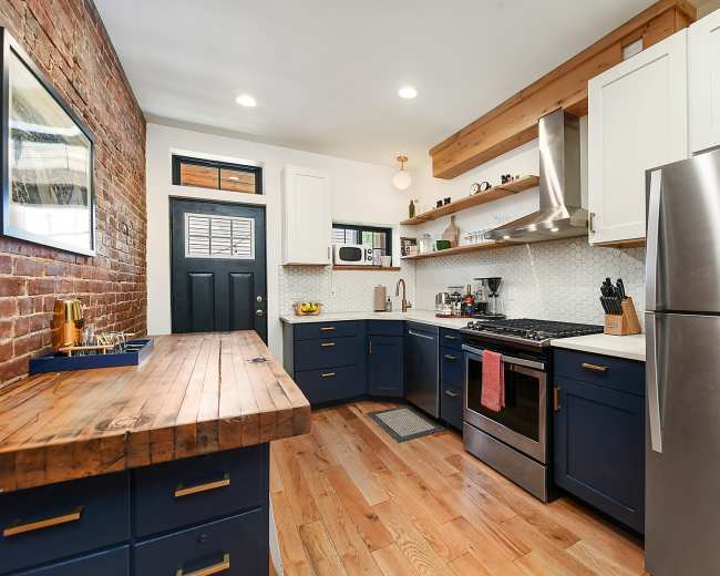 The kitchen features a combination of exposed brick and white cabinetry, with a wooden countertop, stainless steel appliances, and a door leading outside.