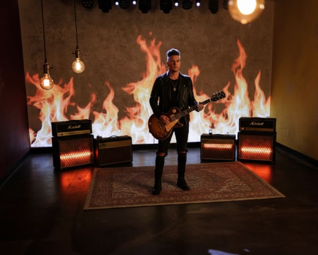 A musician stands on a rug in a dimly lit room with amplifiers and a backdrop of flames.