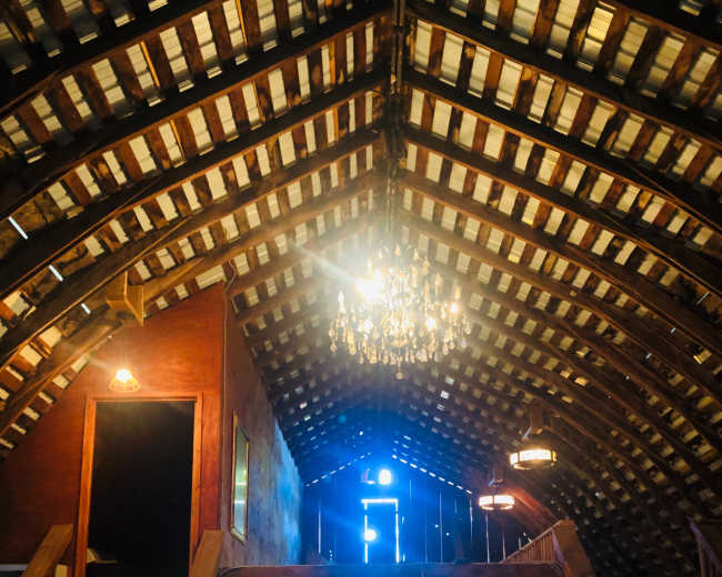 The interior of a barn features a high, wooden ceiling with exposed beams, a chandelier hanging in the center, and stairs leading to an upper level, illuminated by light coming from an open door.