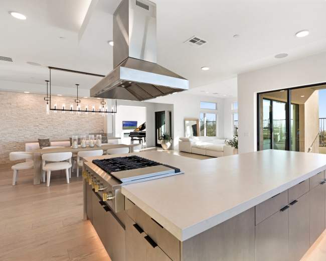 The image shows a modern kitchen with a central island, stainless steel range hood, and an open layout that connects to a dining area and a balcony with a view.