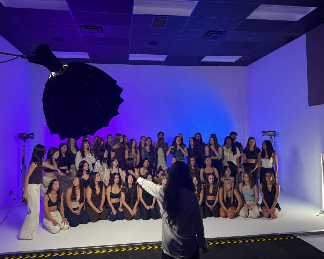 A large group of women is posed for a photoshoot in a studio with colorful lighting and professional equipment.
