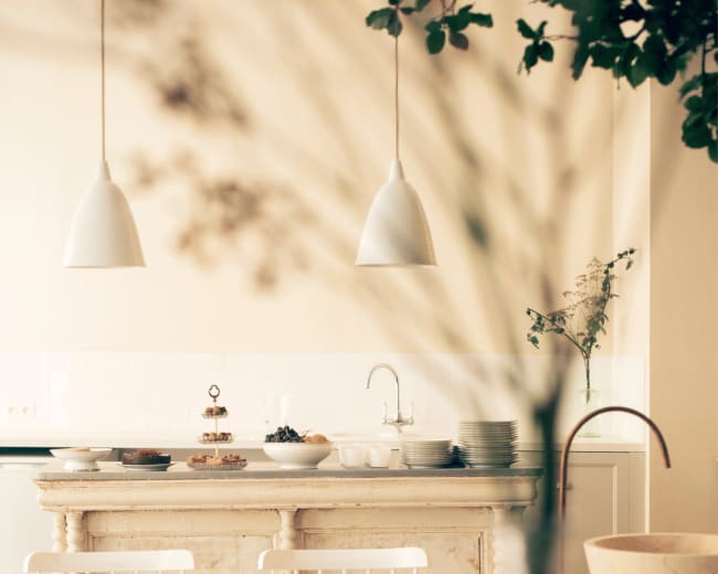 A light-filled kitchen features a rustic island with various plates and a sink, complemented by white chairs and hanging pendant lights.