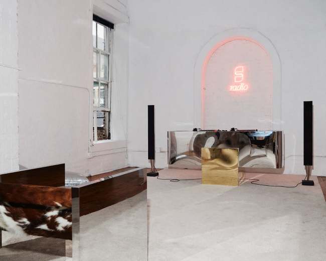 The image depicts a minimalist room featuring a silver and wooden table, a golden object, and a neon sign on the wall.