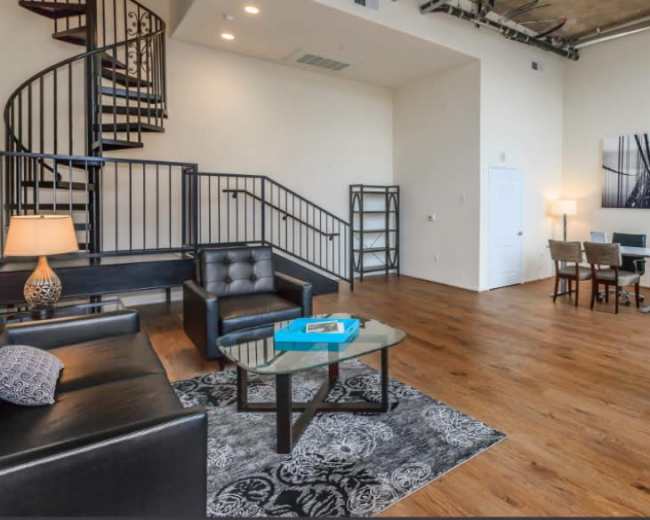 The image shows a modern, two-story loft with a spiral staircase, featuring a living area with seating, a coffee table, and a dining space.