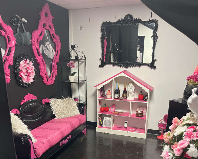 The room features black and pink decor, including a pink sofa, decorative mirrors, and a small shelf displaying bottles and accessories.