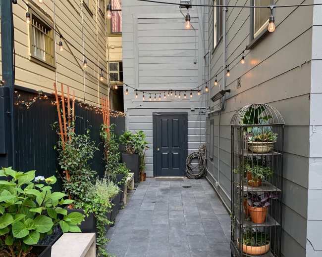 The image shows a narrow courtyard lined with planters, featuring potted plants and string lights overhead, leading to a dark door at the far end.
