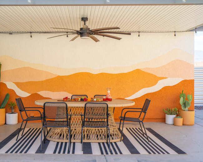 A patio area features a round wicker dining table surrounded by six black chairs, set against a colorful mural of mountains and cacti in pots.