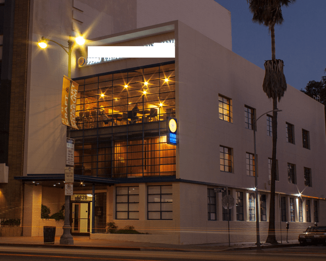 The image shows a modern building with large windows, illuminated at dusk, and featuring a sign that reads "Denny's."