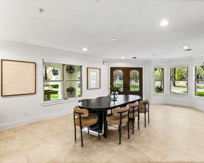 A round table with eight chairs is set in a well-lit room featuring large windows and an entrance door.