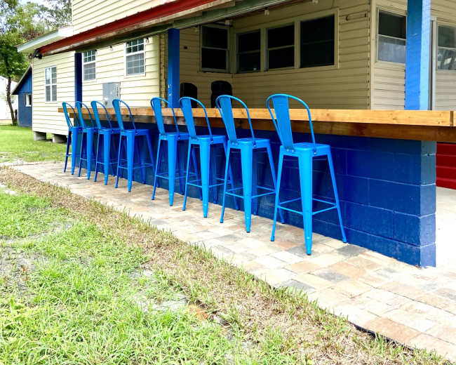 A row of blue metal bar stools is lined up in front of a wooden counter adjacent to a two-story yellow and blue building.