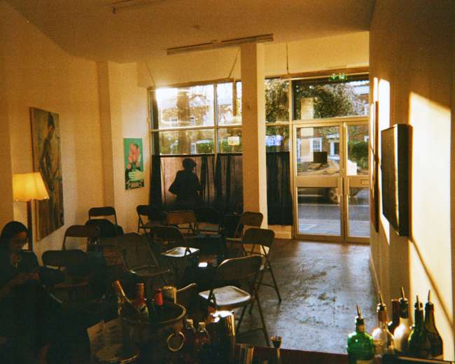 The image shows a sparsely furnished room with empty chairs arranged in rows, a floor lamp, and a person standing by the window.