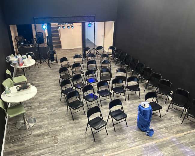 A vacant room arranged with rows of black chairs facing a stage, illuminated by blue lights, with tables and supplies in the background.