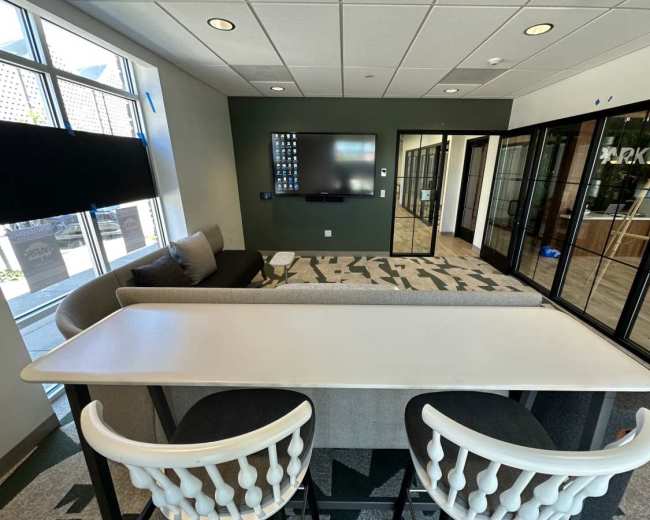 The image shows a modern office space featuring a long table with two chairs, a sofa, a television on the wall, and glass-paneled meeting rooms.