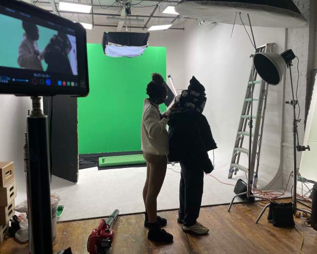 A person is adjusting clothing on another individual in a studio setting with a green screen and various lighting equipment.