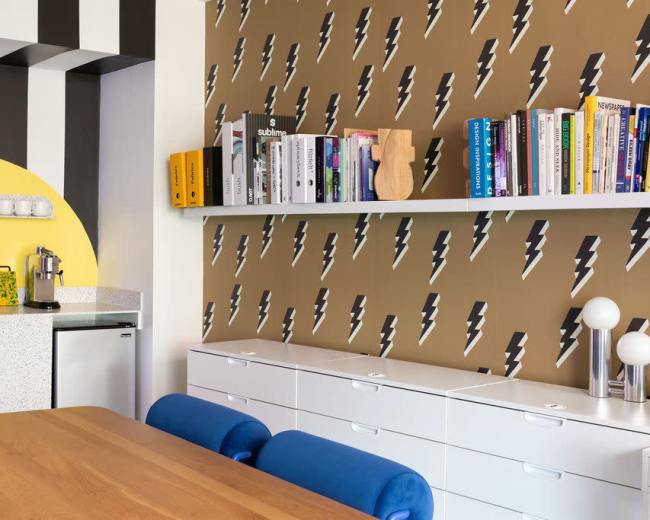 The image shows a modern kitchen with a patterned wall featuring lightning bolts, a wooden dining table, and a white shelf filled with books.