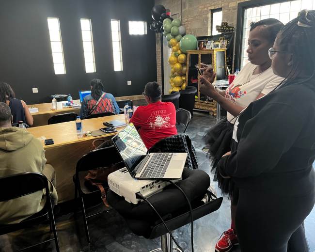 A group of people sits at a long table in a modern room while two individuals stand beside a projector and laptop, engaging with a presentation.