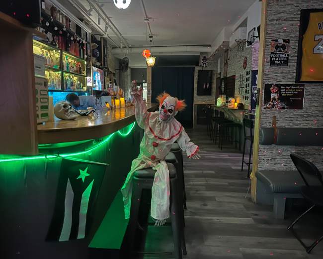 A clown mannequin sits on a bar stool in a dimly lit bar decorated with various memorabilia.