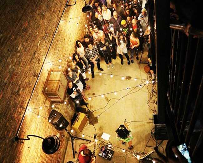 A crowd gathers in a dimly lit venue with exposed brick walls, watching a performer on stage surrounded by string lights.