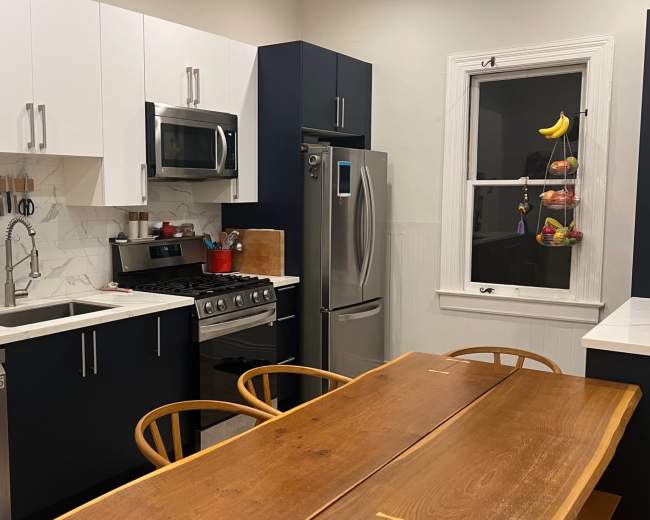 The image shows a modern kitchen with blue and white cabinetry, stainless steel appliances, and a wooden dining table.