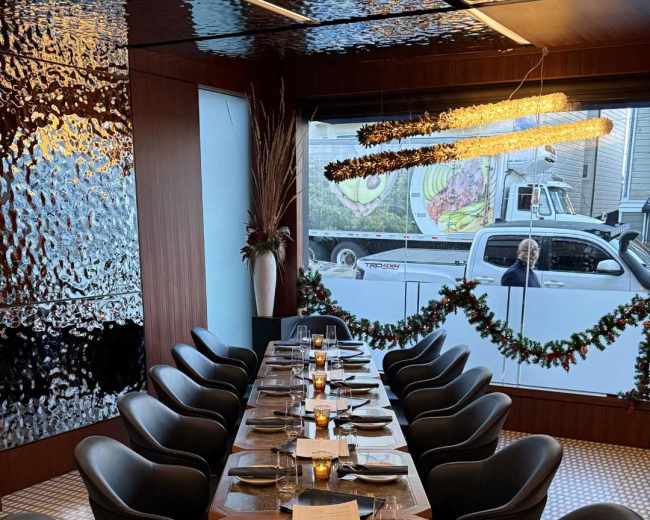A long dining table with black chairs is set for a meal in a modern restaurant, featuring a decorative ceiling and window view of an outside street.