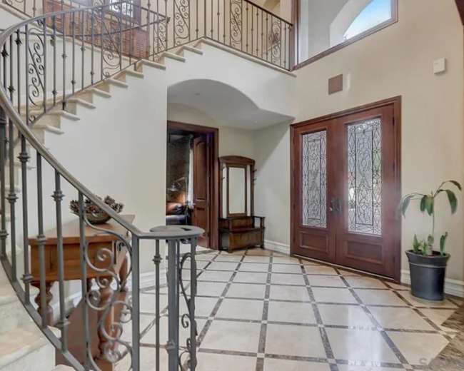 The image shows a foyer with a curved staircase, decorative iron railing, and double doors with glass panels opening into the space.