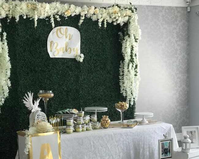A decorated event space with a green floral backdrop, a table covered with treats, and decorative blocks labeled "A," "B," and "C" in front of the table.