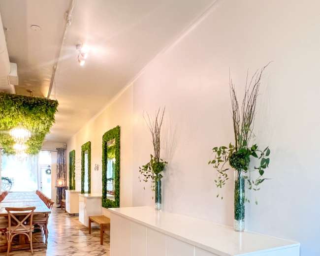 The image shows a hallway with white walls, a marble floor, and a long white cabinet, accented by vases of greenery and framed with vertical plant installations.