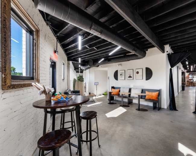 The image shows a modern, open interior space with black exposed beams, seating areas, and a small table arranged near a window.