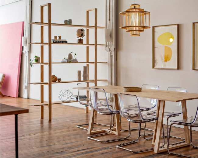A modern dining area features a wooden table, clear acrylic chairs, a tall wooden shelf, and framed artworks on the wall.