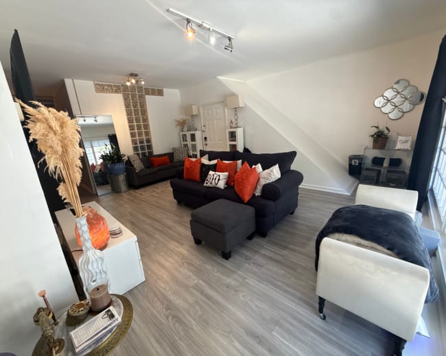 The image shows a spacious living room featuring a mix of dark and light furniture, with orange decorative pillows and an area rug contrasting the light wood flooring.