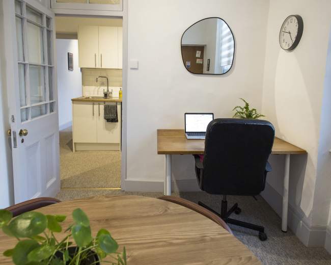 The image shows a small home office setup featuring a desk with a laptop, a chair, and a mirror, with a kitchen area visible in the background.