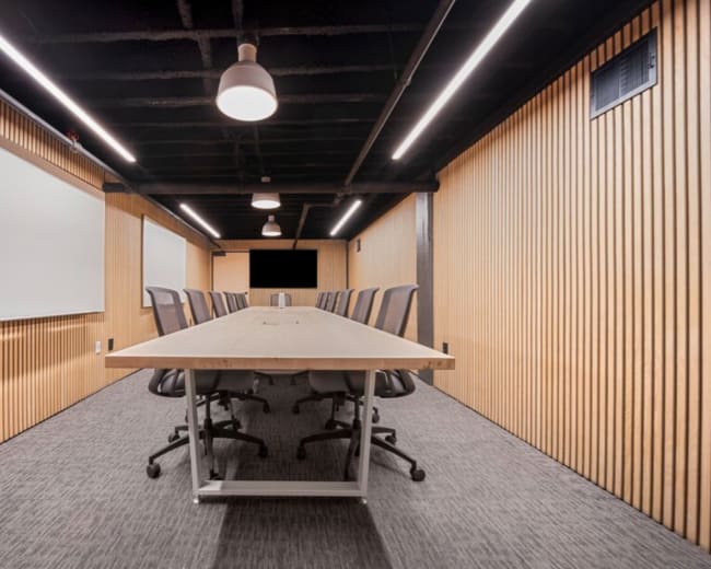 A spacious conference room features a long wooden table surrounded by ergonomic chairs, with wooden paneling on the walls and whiteboards on either side.