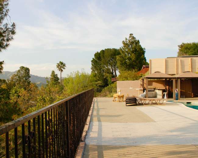 A wooden railing overlooks a patio area with lounge chairs and an outdoor space beside a swimming pool, surrounded by trees and hills.