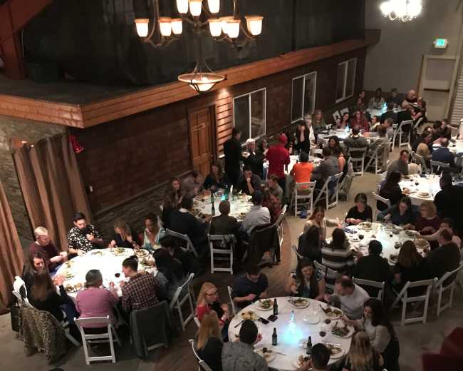 A large group of people is gathered around several round tables in a dimly lit banquet hall, enjoying a meal together.