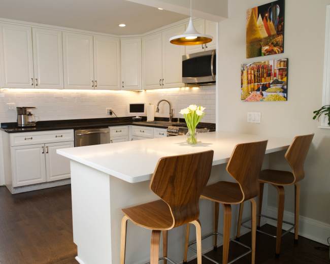 A modern kitchen features white cabinetry, a black countertop, and a breakfast bar with four wooden stools.
