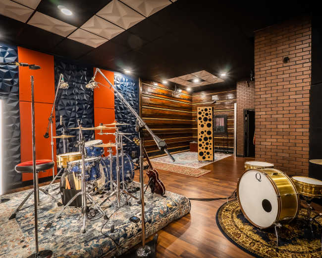A music recording studio featuring a drum set on a patterned rug, surrounded by soundproofed walls and acoustic panels.