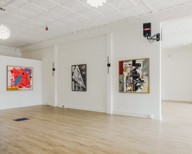The image shows a spacious, well-lit gallery with white walls displaying several abstract paintings and wooden flooring.