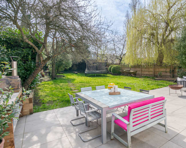 A patio with a pink-accented dining table and chairs overlooks a lawn bordered by trees and shrubs.