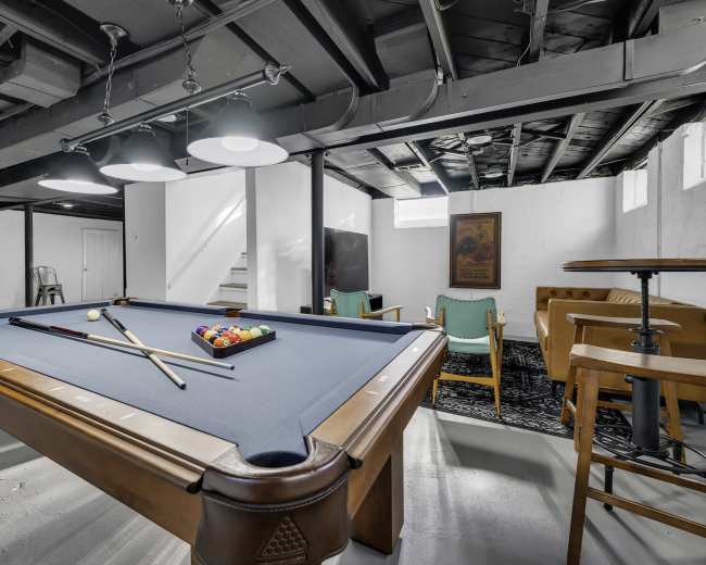 A pool table with cues and balls is centered in a basement, surrounded by seating areas and a television.